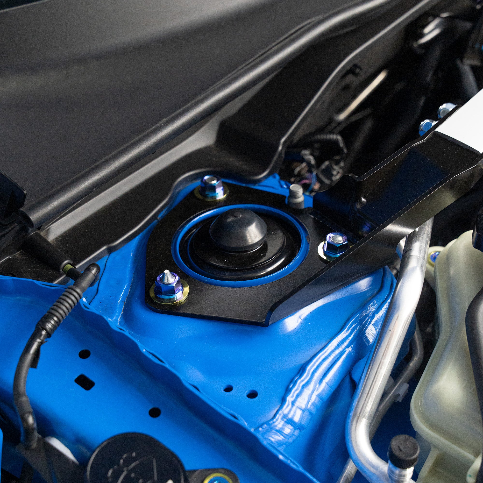 Close-up of a blue car engine bay with titanium strut tower nuts and washers