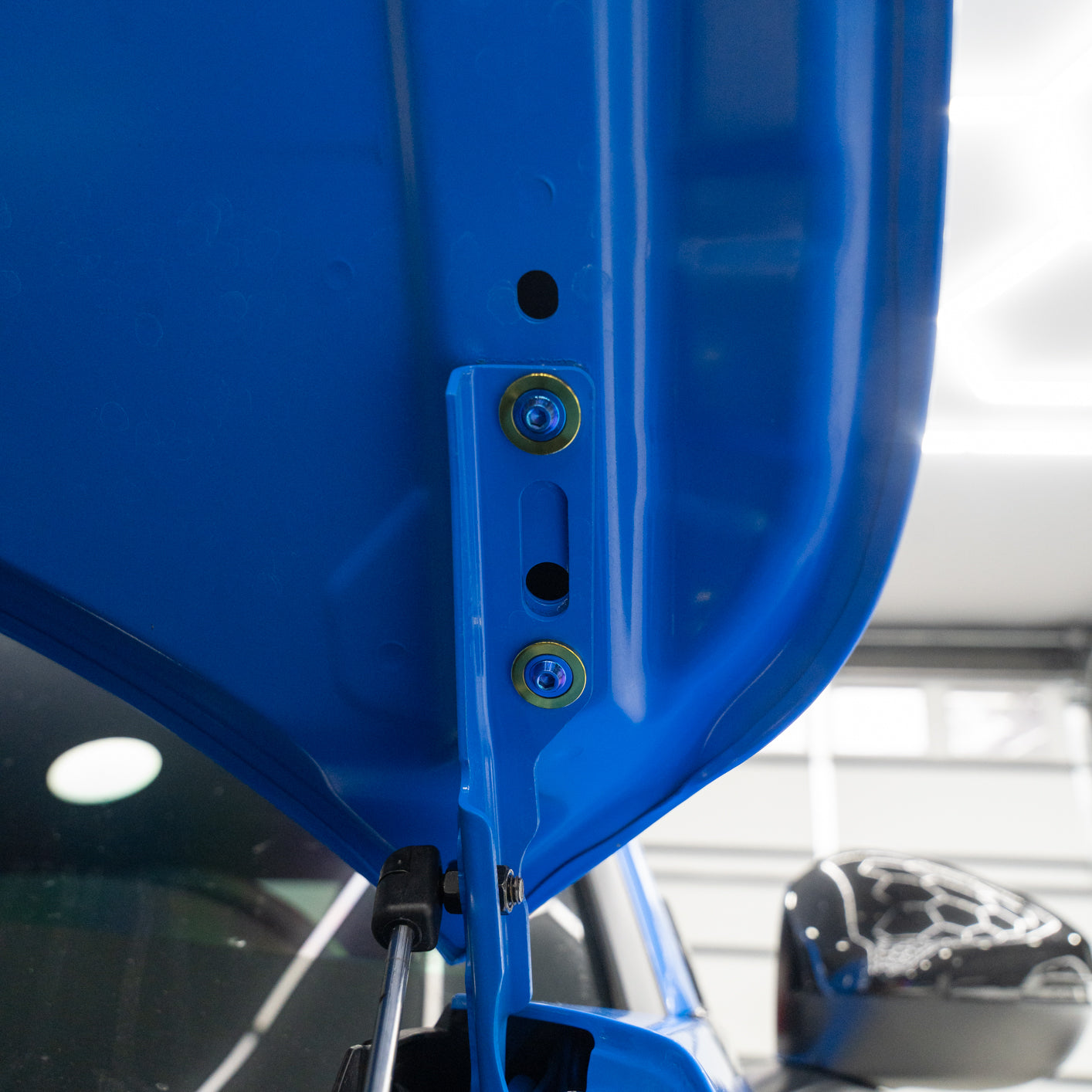 Close-up of a blue car hood hinge with titanium hardware.