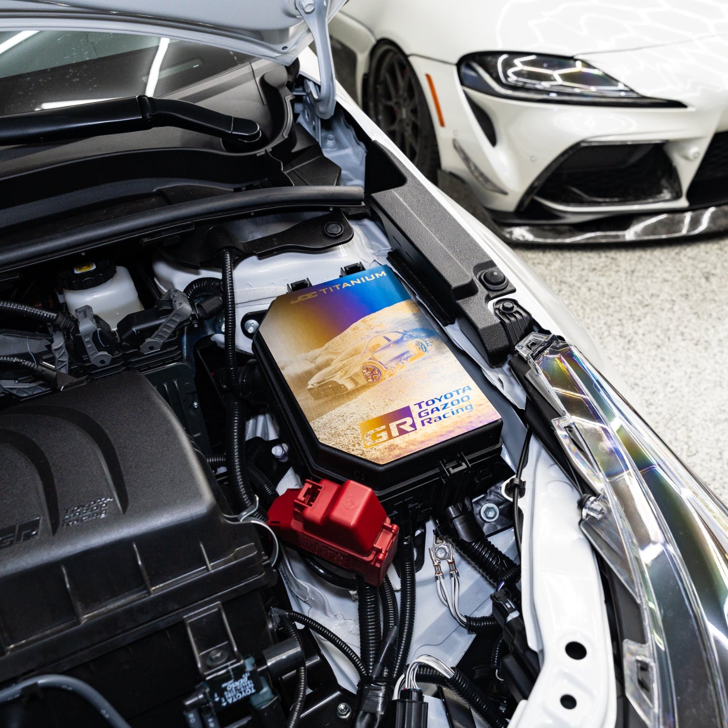Car engine displaying a Toyota GR Corolla engine bay fuse box cover with a white Supra in the background.