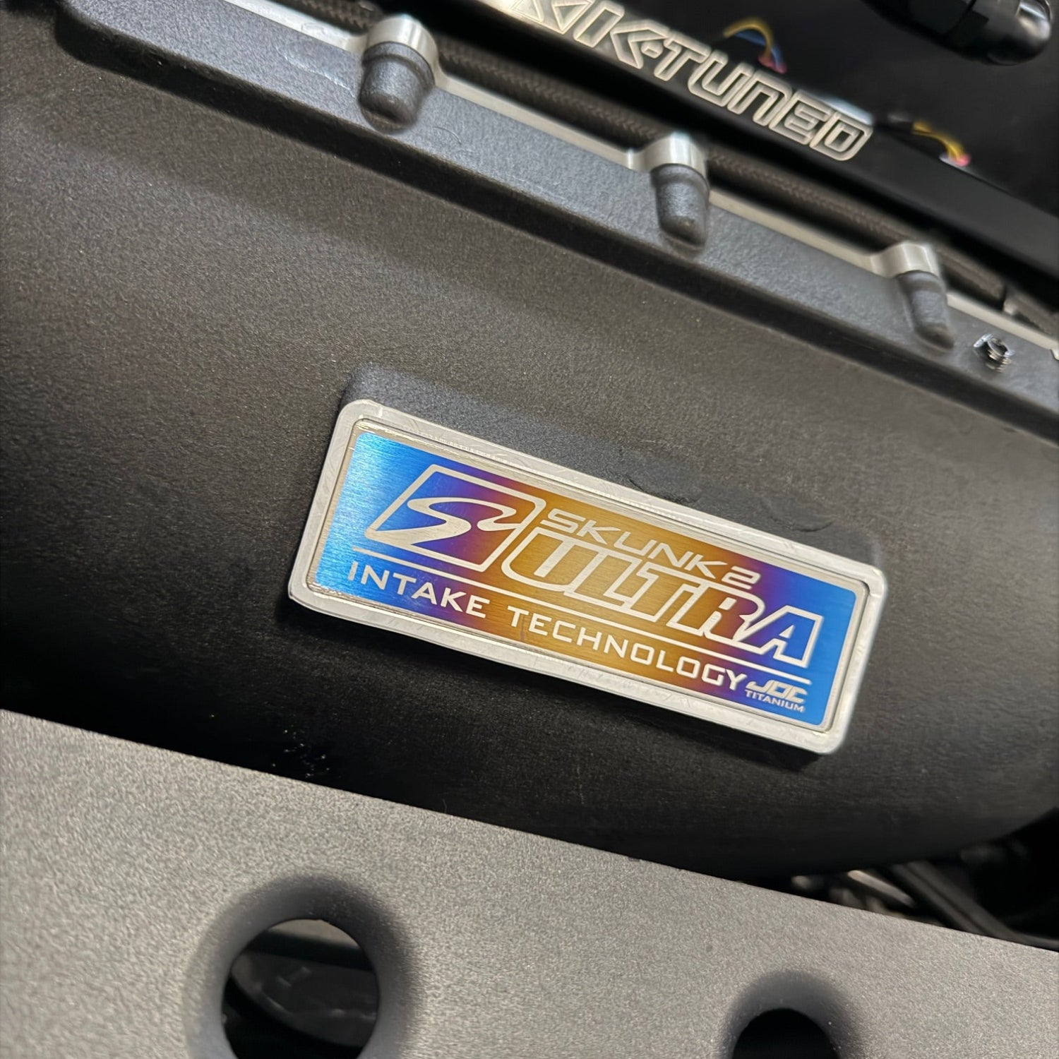 Close up of the JDC Titanium Skunk2 intake manifold badge installed on a black intake manifold.