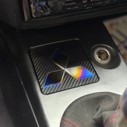 A close-up of a car center console featuring a square carbon fiber ashtray delete panel with a brushed, burnt titanium Mitsubishi logo decal. A Kenwood radio is mounted above, and a gear shift knob is visible in the foreground.
