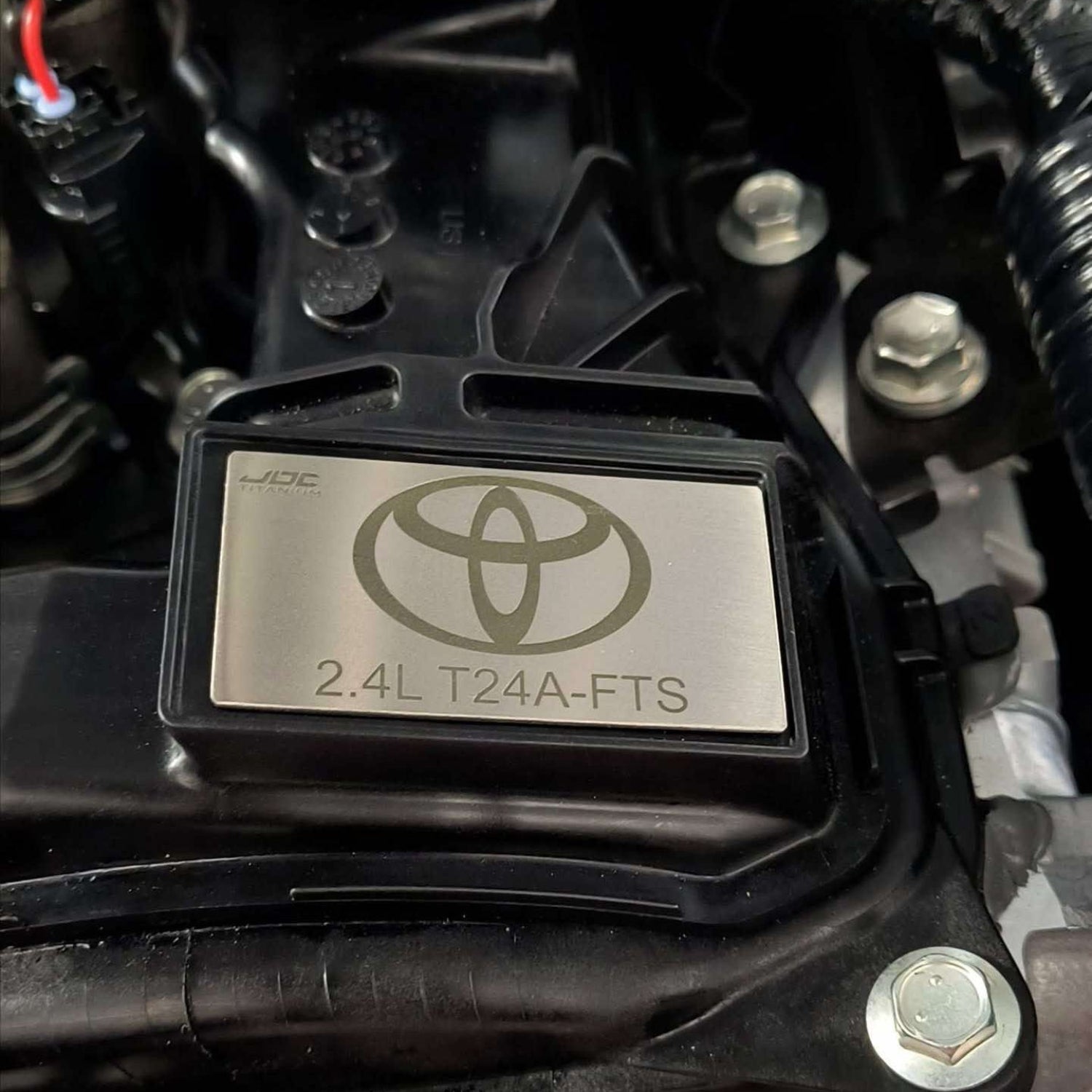 JDC Titanium badge on a Toyota Tacoma valve cover with '2.4L T24A-FTS' label in an engine bay.